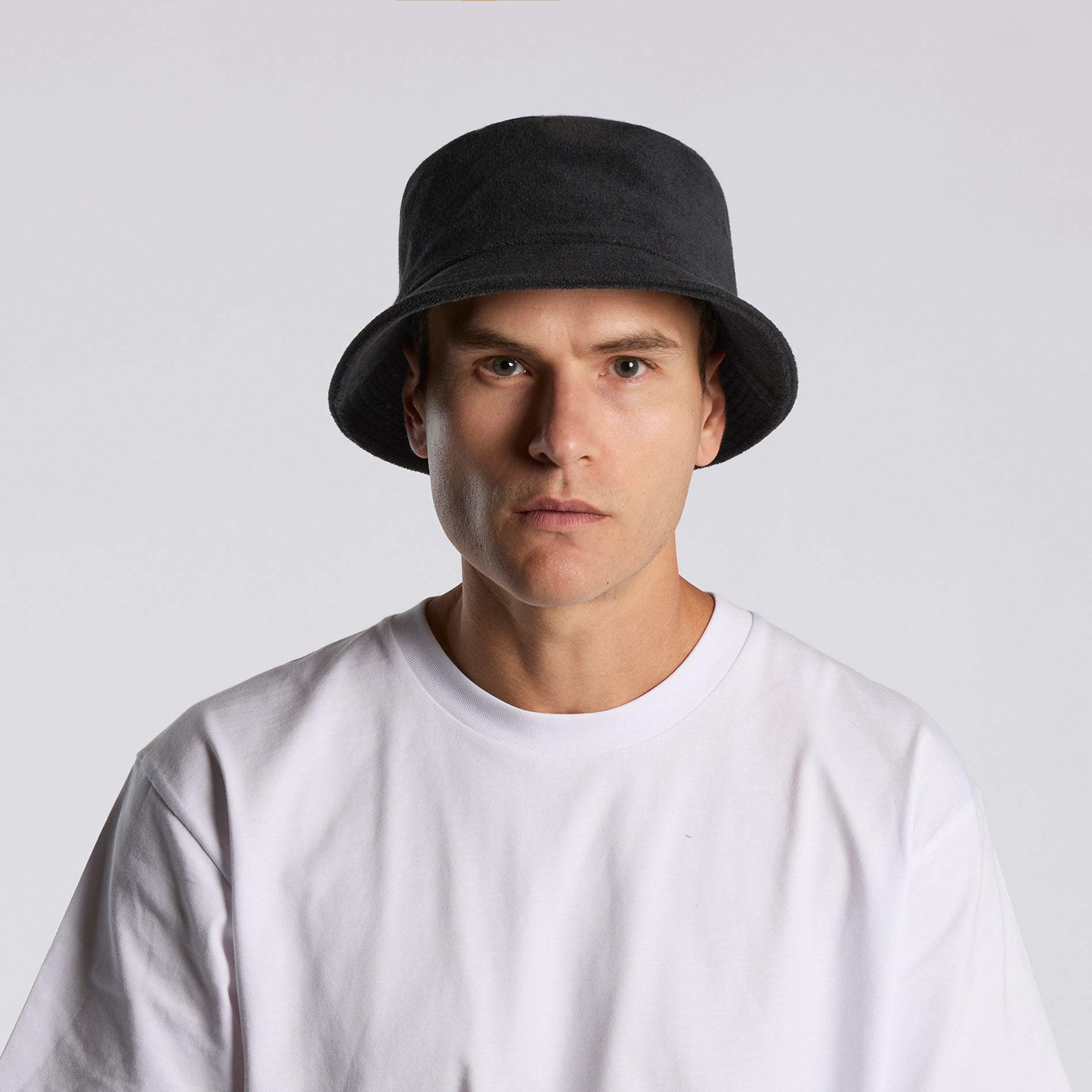 Man wearing a black AS Colour terry bucket hat and white t-shirt on a light gray background