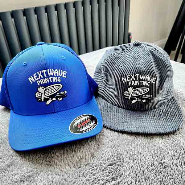 Two baseball caps, one blue and one gray, with 'NextWave Printing' embroidered logo on a textured surface.