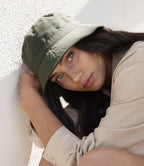 Person wearing a green cap and beige shirt against a textured white wall