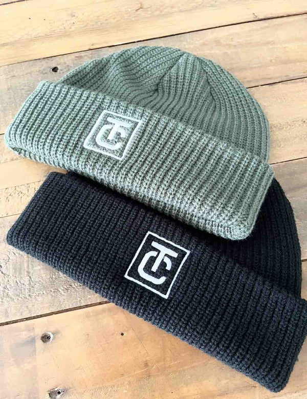 Two knit beanie hats, one green and one black, with an embroidered logo on a wooden surface.