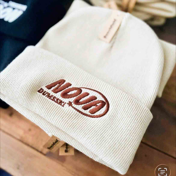Beige knit beanie with embroidered brown logo on a wooden surface