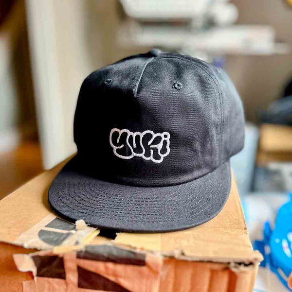 Black cap with 'YUKI' embroidered logo on a cardboard box