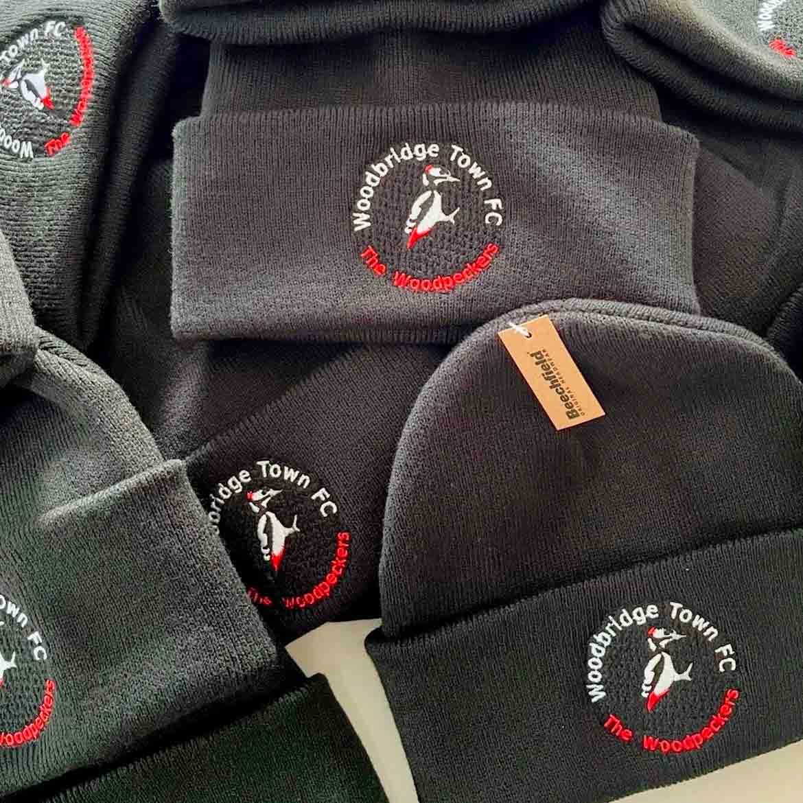 Collection of black beanies with Woodbridge Town FC embroidered logo on a beige surface