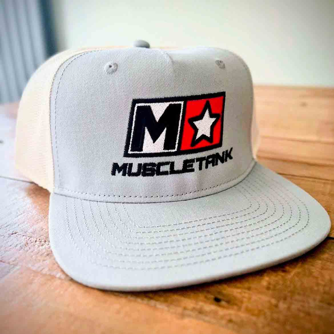 Gray trucker style cap with embroidered Muscle Tank logo on a wooden surface