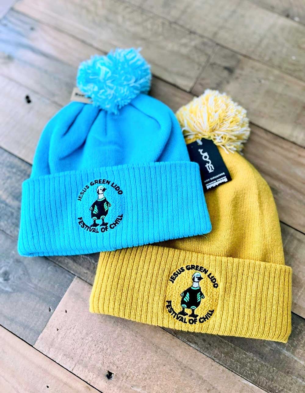 Two beanies, one blue and one yellow, with pom-poms with an embroidered logo on a wooden surface.