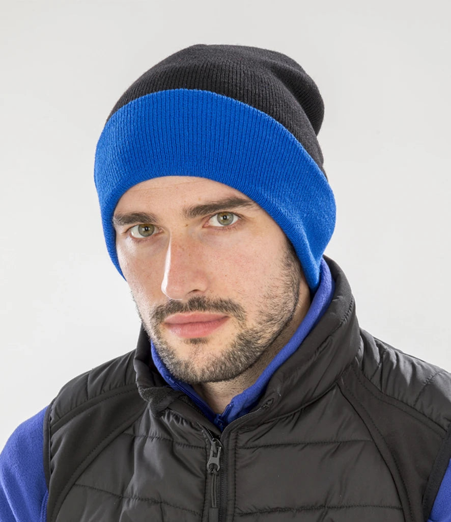 male model wearing Result Recycled Compass Beanie in colour black and royal blue photographed on a grey backrground
