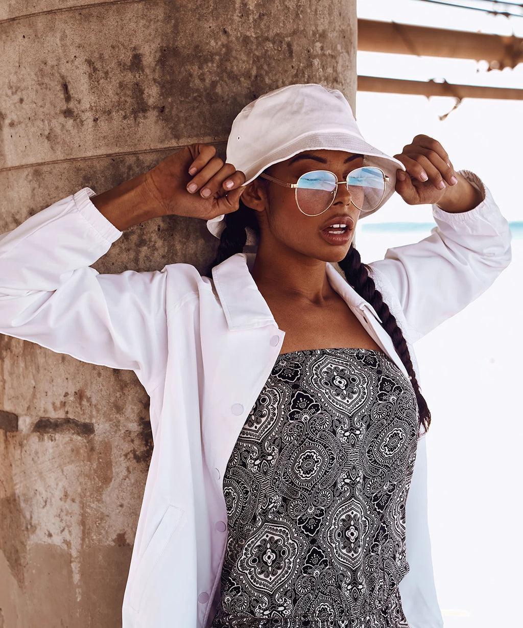 Woman wearing white flexfit cotton twill bucket hat  and sunglasses, standing against a textured wall.