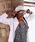 Woman wearing white flexfit cotton twill bucket hat  and sunglasses, standing against a textured wall.