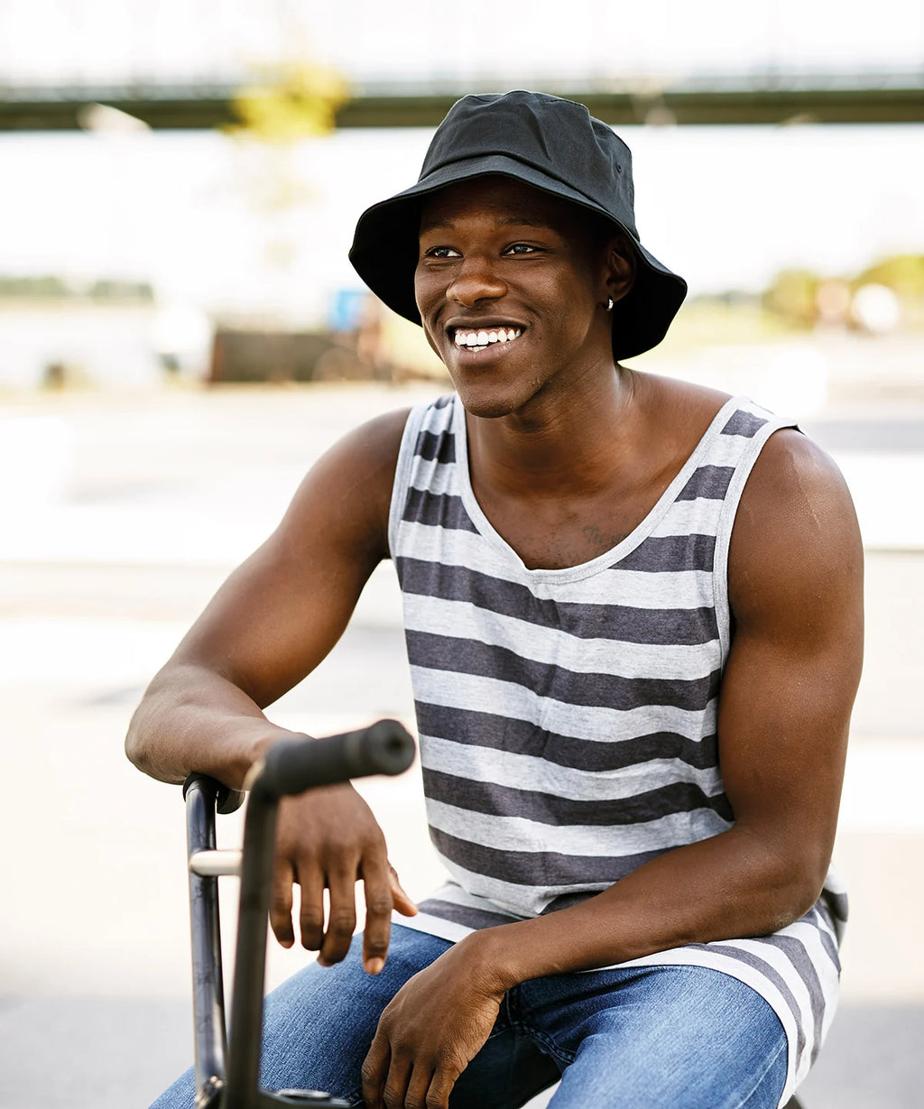 Man wearing a black and white striped tank top, blue jeans and black flexfit cotton twill bucket hat sitting outdoors.