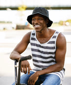 Man wearing a black and white striped tank top, blue jeans and black flexfit cotton twill bucket hat sitting outdoors.